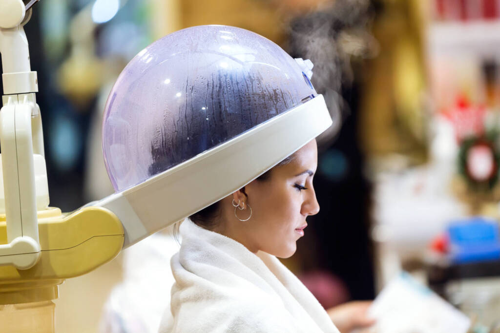 Beautiful young woman drying hair under machine in a beauty salo