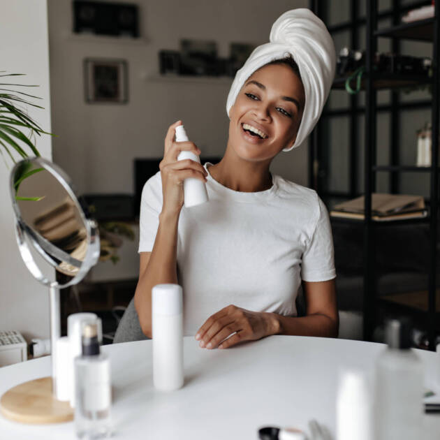 Brown-eyed charming woman in white t-shirt and towel on head spread spray at face. Cheerful lady sm